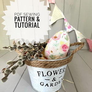 May include: A white metal bucket with a brown rope handle and the text "FLOWER & GARDEN" printed on the side. The bucket contains a floral patterned fabric egg and a sprig of pussy willow.