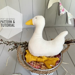 Può includere: Un'oca di stoffa bianca con un becco giallo seduta in un secchio di metallo con un manico di corda. Il secchio ha la scritta "FLOWER GARDEN" stampata sul lato. L'oca è circondata da carta triturata rosa e marrone.