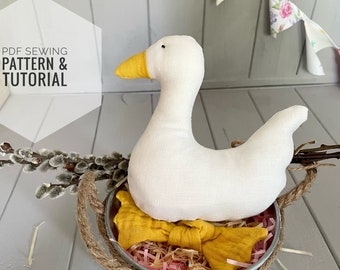 Puede incluir: Un ganso de tela blanca con un pico amarillo sentado en un cubo de metal con un asa de cuerda. El cubo tiene las palabras "FLOWER GARDEN" impresas en el lateral. El ganso está rodeado de papel triturado rosa y marrón.