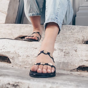 May include: A woman wearing blue jeans and black sandals with gold accents sits on a set of concrete steps in front of a gray door.