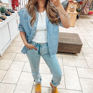 Puede incluir: Una mujer con una camisa de botones a rayas azul claro sobre un top blanco sin mangas, jeans azul claro y sandalias amarillas.
