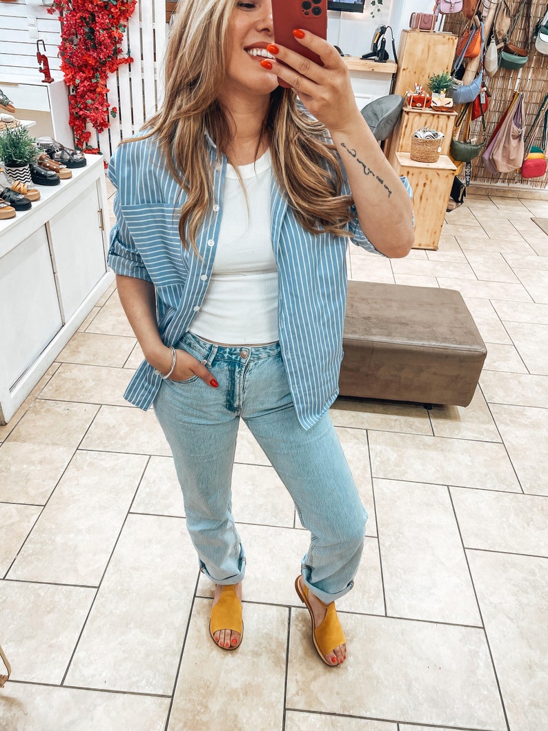 Puede incluir: Una mujer con una camisa de botones a rayas azul claro sobre un top blanco sin mangas, jeans azul claro y sandalias amarillas.
