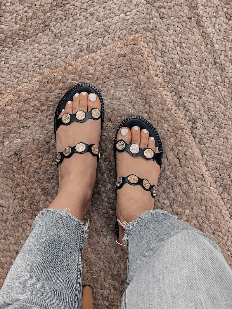 May include: Black leather sandals with gold-toned round embellishments. The sandals have two straps across the foot and a simple design.