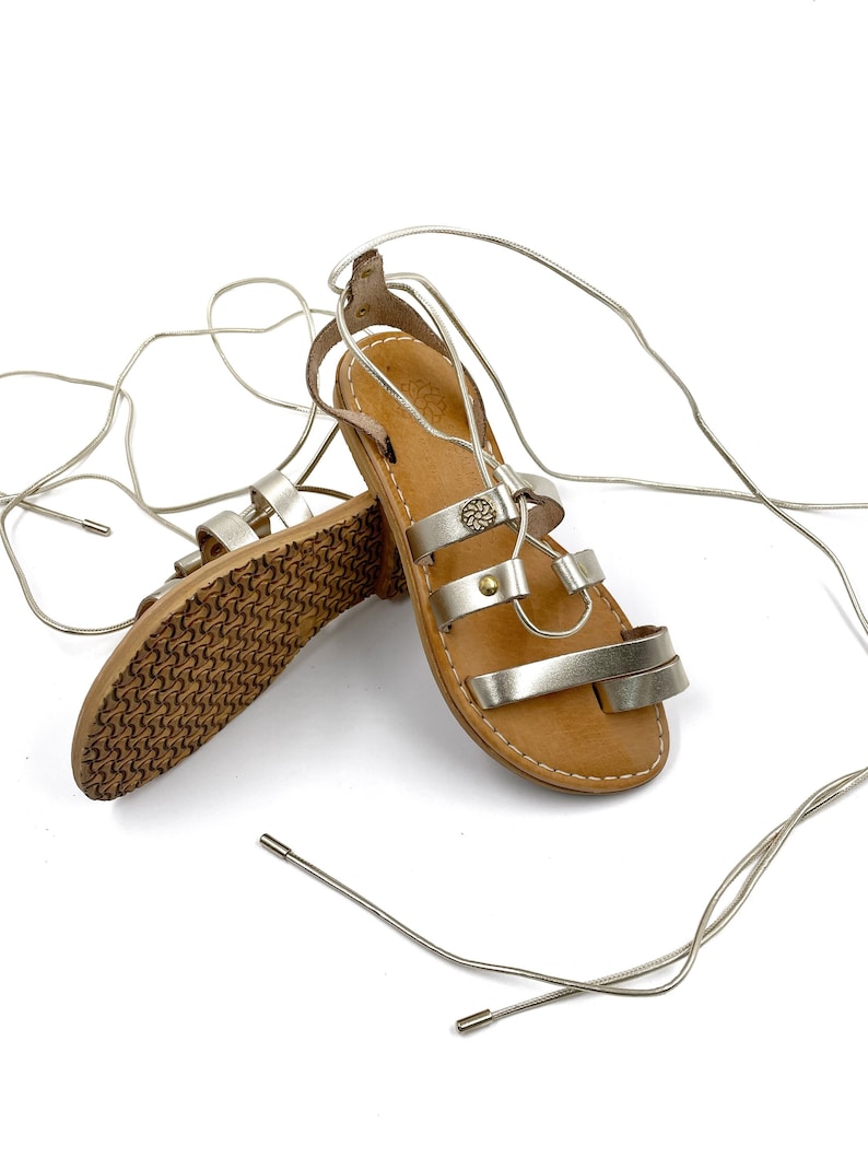 May include: Gold leather sandals with a lace-up design. The sandals have a brown leather sole and a gold metal buckle.