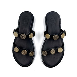 May include: A pair of black leather sandals with gold-toned metal accents. The sandals have two straps across the foot and a rounded toe.