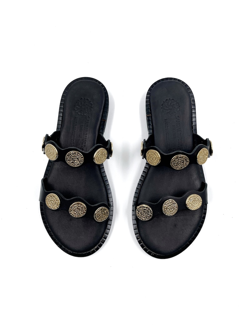 May include: A pair of black leather sandals with gold-toned metal accents. The sandals have two straps across the foot and a rounded toe.