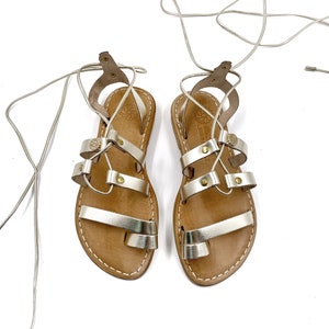May include: A pair of gold leather sandals with lace-up closures. The sandals have a brown leather sole and are tied with a thin gold cord.