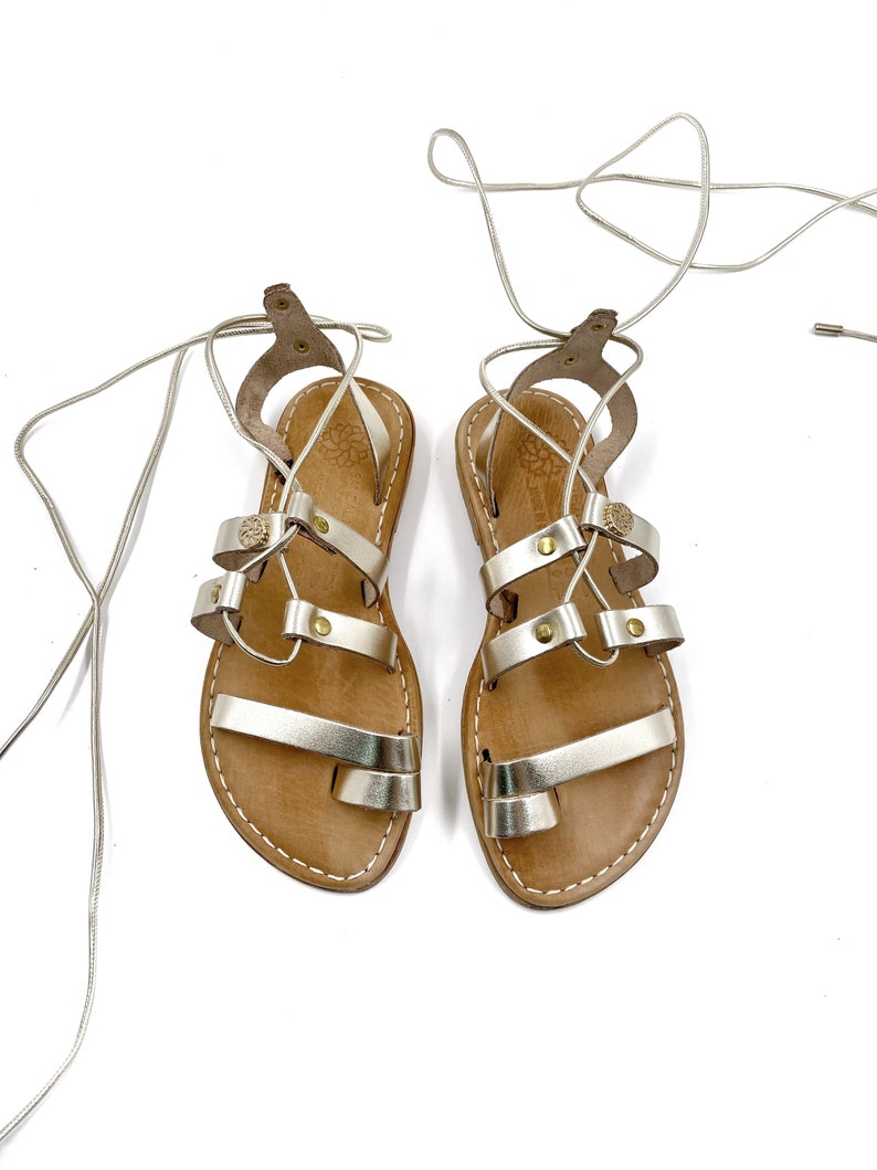 May include: A pair of gold leather sandals with lace-up closures. The sandals have a brown leather sole and are tied with a thin gold cord.