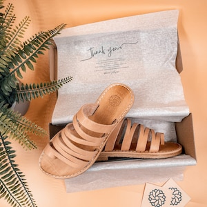 May include: Tan leather sandals with multiple straps, packaged in a white tissue paper-lined box with a thank you note. The box is sitting on a light brown surface. Two small business cards with a floral logo and the text "SANDAL MANIA" are visible in the bottom right corner.