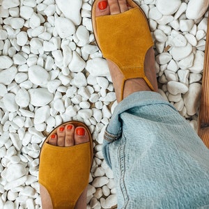 Puede incluir: Un par de sandalias de ante amarillo mostaza con una correa alrededor del tobillo. Las sandalias est&aacute;n sobre un fondo de guijarros blancos.