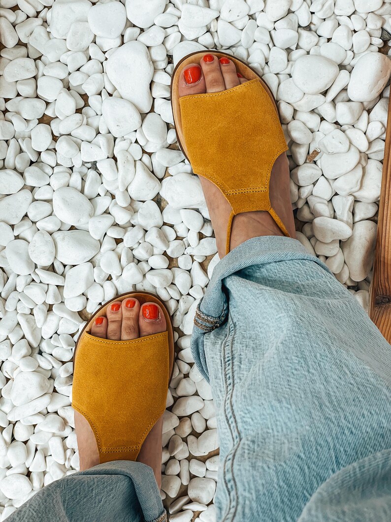 Puede incluir: Un par de sandalias de ante amarillo mostaza con una correa alrededor del tobillo. Las sandalias est&aacute;n sobre un fondo de guijarros blancos.