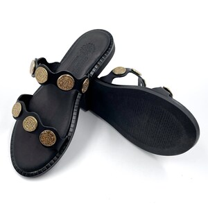 May include: Black leather sandals with gold-toned round embellishments. The sandals have a simple design and a flat sole.