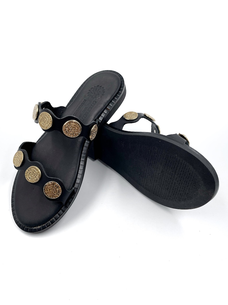 May include: Black leather sandals with gold-toned round embellishments. The sandals have a simple design and a flat sole.