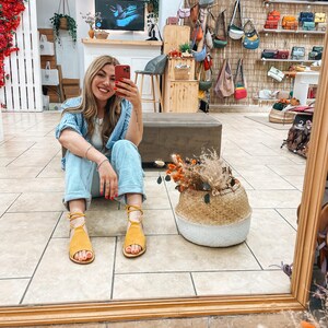 Puede incluir: Una mujer con una camisa a rayas azul y blanca y pantalones vaqueros est&aacute; sentada frente a un espejo. Est&aacute; sosteniendo un tel&eacute;fono en su mano derecha y sonriendo. Lleva sandalias amarillas. Hay una cesta de mimbre con flores secas delante de ella. El espejo refleja una tienda con un letrero que dice "AIAM JADIA".