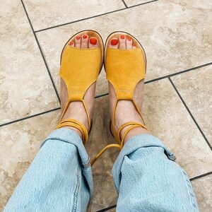 Puede incluir: Un par de sandalias de ante amarillo con ataduras en el tobillo. Las sandalias tienen un dise&ntilde;o sencillo y se llevan con vaqueros azul claro.