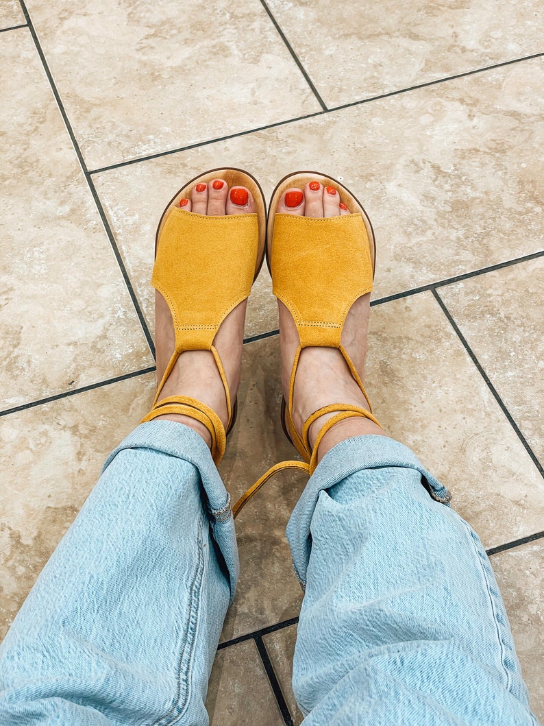 Puede incluir: Un par de sandalias de ante amarillo con ataduras en el tobillo. Las sandalias tienen un dise&ntilde;o sencillo y se llevan con vaqueros azul claro.