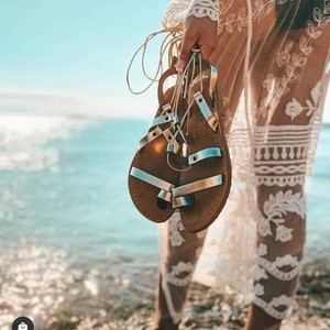 May include: A pair of brown leather sandals with silver straps. The sandals are tied together with a gold cord.