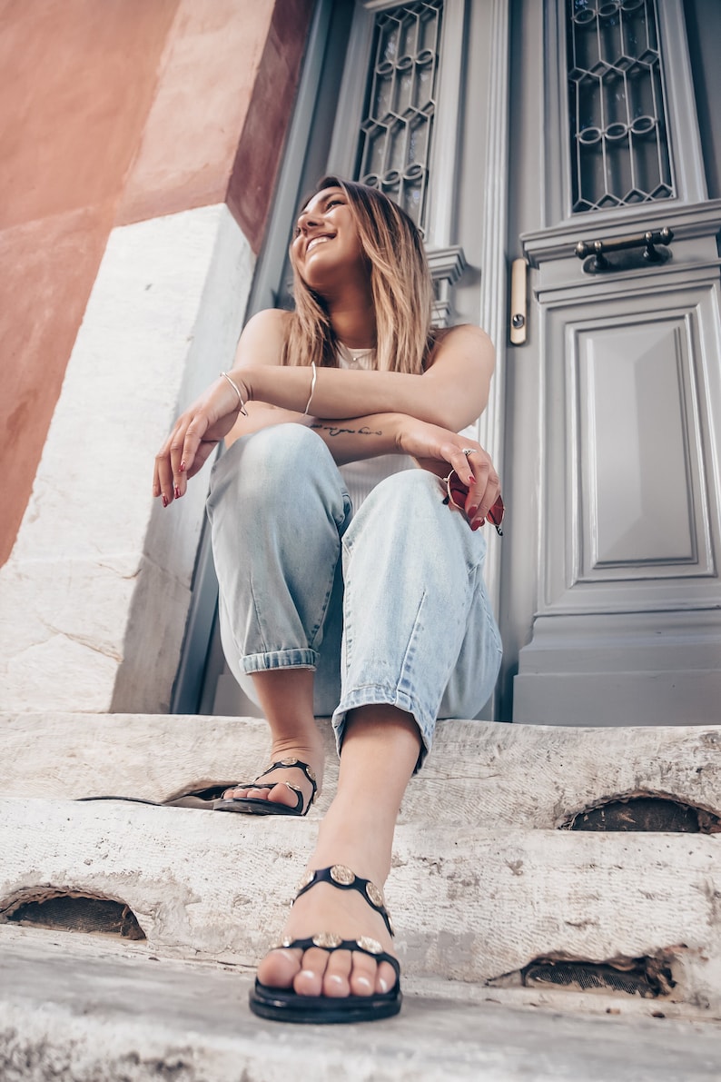 May include: A woman wearing blue jeans and black sandals with gold accents sits on a set of concrete steps in front of a gray door.