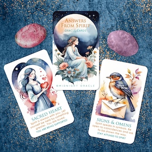 May include: Oracle cards with watercolor illustrations and text. One card reads "ANSWERS FROM SPIRIT ORACLE CARDS." Another card says "SACRED HEART" with supporting text. A third card reads "SIGNS & OMENS" with supporting text. Two pink stones are also present.