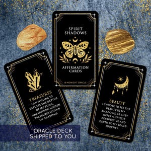 May include: Black oracle cards with gold accents and text. One card reads "Spirit Shadows Affirmation Cards" with a butterfly design. Another card says "Treasures" and the third card says "Beauty". Two gold-colored stones are also in the image.