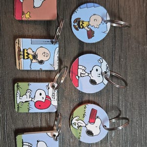 May include: A collection of keyrings featuring Snoopy and Charlie Brown characters. The keyrings have silver rings and are rectangular and circular. The images on the keyrings are from the Peanuts comic strip.
