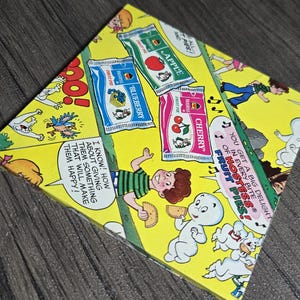 以下が含まれることがあります： 漫画をテーマにした黄色の箱で、漫画のキャラクターとフルーツパイのパッケージが描かれています。箱には「Apple」、「Blueberry」、「Cherry」の文字と「Fruit Pies！」というフレーズが表示されています。