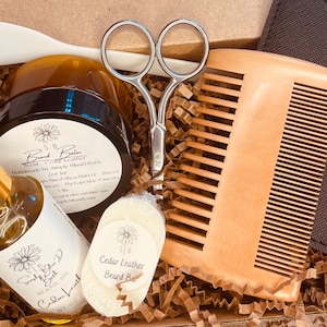 May include: A gift box filled with beard care products, including a beard balm, beard oil, beard bar, wooden comb, and a pair of scissors. The products are labeled with the text "Cedar Leather Beard Balm", "Cedar Leather Beard Oil", and "Cedar Leather Beard Bar".
