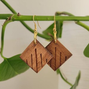 May include: A pair of wooden earrings with a diamond shape and a wood grain pattern. The earrings are made of dark brown wood and have gold hooks.