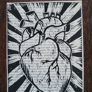 May include: A black and white linocut print of a human heart with a radiating sunburst pattern behind it. The heart is surrounded by text from a book.