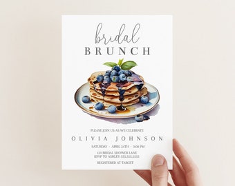Puede incluir: Una tarjeta de invitación blanca con el texto "bridal brunch" en una fuente de escritura. La tarjeta presenta una ilustración de acuarela de una pila de panqueques cubiertos con arándanos y jarabe.