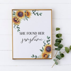 May include: A framed print with a white background and gold frame. The print features a watercolor illustration of sunflowers and greenery with the text "She found her sunshine".