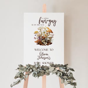 May include: A white sign on a wooden easel with the text "A little fun-guy is on the way." A floral illustration of mushrooms and flowers is centered on the sign. The sign reads "Welcome to Olivia Johnson's Baby Shower."