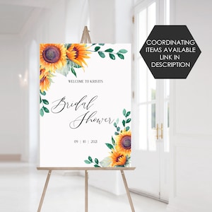 May include: A white welcome sign with a sunflower and greenery design. The text reads "Welcome to Kristi's Bridal Shower 09 | 10 | 2021".