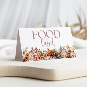 Może przedstawiać: Biała, złożona etykieta z napisem "FOOD label" w kolorze mauve. Dolna krawędź ozdobiona jest akwarelowymi kwiatami w odcieniach pomarańczu, brązu i zieleni. Etykieta jest wyświetlana na drewnianej powierzchni.