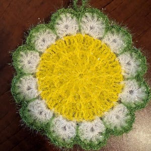 May include: A handmade, crocheted flower-shaped item with a yellow center, white petals, and a green, glittery edge. The item has a loop for hanging and is likely a decorative or functional item, such as a coaster or washcloth.