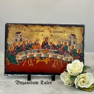 May include: A rectangular religious icon depicting the Last Supper, with figures seated around a table. The artwork has a gold background and architectural details. The icon is mounted on a dark, textured stone base, with the text "Byzantium Tales" at the bottom.