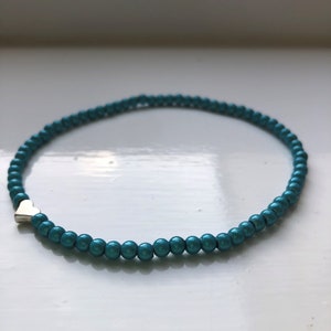 Può includere: Un braccialetto di perline blu turchese con un ciondolo a forma di cuore argentato.