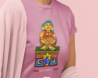 May include: A woman wearing a pink t-shirt with a colorful graphic of a seated figure and the text "XOCHIMILLI". She is also wearing a pink cardigan and a pink skirt. She is holding a white rose.