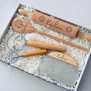 May include: A wooden baking set in a white box, including a rolling pin, spoon, dough cutter, and pastry blender. Each wooden item is engraved with a cookie graphic and the name "Kailani". The set is nestled in white paper filler.