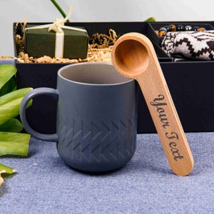 Puede incluir: Una taza de cer&aacute;mica gris con un patr&oacute;n geom&eacute;trico y una cuchara de madera con el texto "Your Text" grabado en ella.