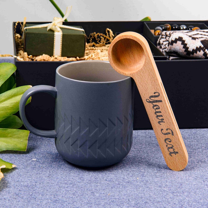 Puede incluir: Una taza de cer&aacute;mica gris con un patr&oacute;n geom&eacute;trico y una cuchara de madera con el texto "Your Text" grabado en ella.