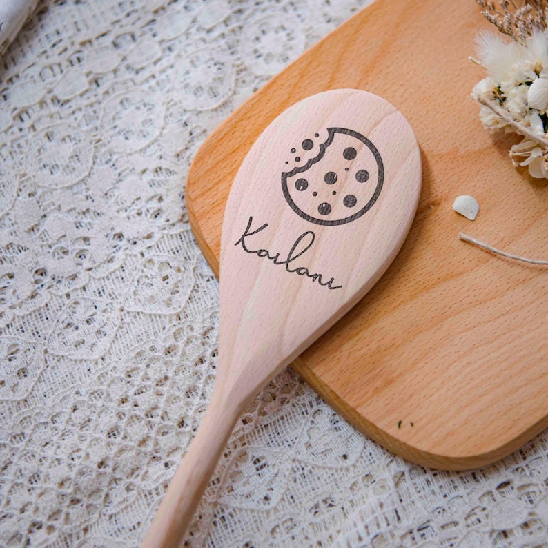 May include: A wooden spoon with a cookie graphic and the name "Karlane" engraved on the handle. The spoon is light brown and rests on a wooden cutting board. The cookie graphic is a circle with a bite taken out of it.