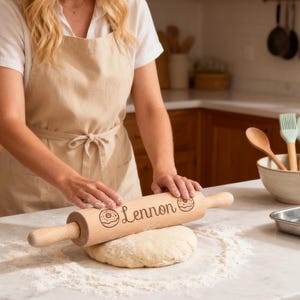 May include: A wooden rolling pin with the name "Lennon" and a donut graphic is used to roll out dough on a white countertop. The rolling pin is light brown, and the dough is surrounded by flour. The scene takes place in a kitchen.
