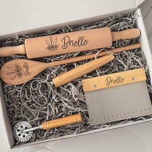 Puede incluir: Un conjunto de utensilios de cocina de madera, que incluye un rodillo y una cuchara, presentados en una caja blanca con relleno de papel. Cada herramienta está grabada con el nombre "Brielle" y diseños florales. La rasqueta para masa tiene una hoja metálica con medidas en cm.