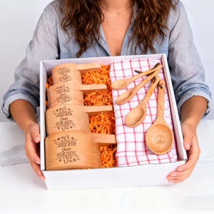 May include: A gift box containing wooden measuring cups and spoons. The cups are engraved with the name "Josie" and floral designs. The box also includes a red and white checkered cloth and orange packing material.
