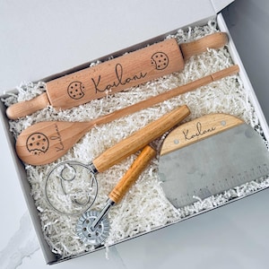 May include: A wooden baking set in a white box, including a rolling pin, spoon, dough scraper, and a whisk. Each wooden item is engraved with a cookie graphic and the name "Kailani". The set is nestled in white paper filler.