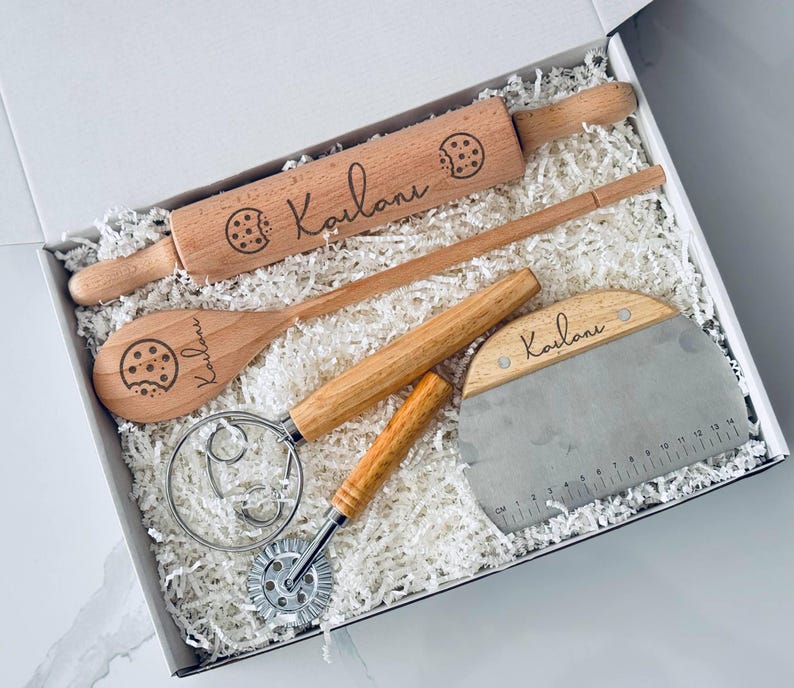 May include: A wooden baking set in a white box, including a rolling pin, spoon, dough scraper, and a whisk. Each wooden item is engraved with a cookie graphic and the name "Kailani". The set is nestled in white paper filler.