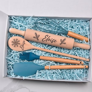 May include: A gift box with blue shredded paper, holding a wooden rolling pin, whisk, spoon, brush, and spatula. The rolling pin and spoon are engraved with the name "Eloise" and a snowflake or pine branch design.
