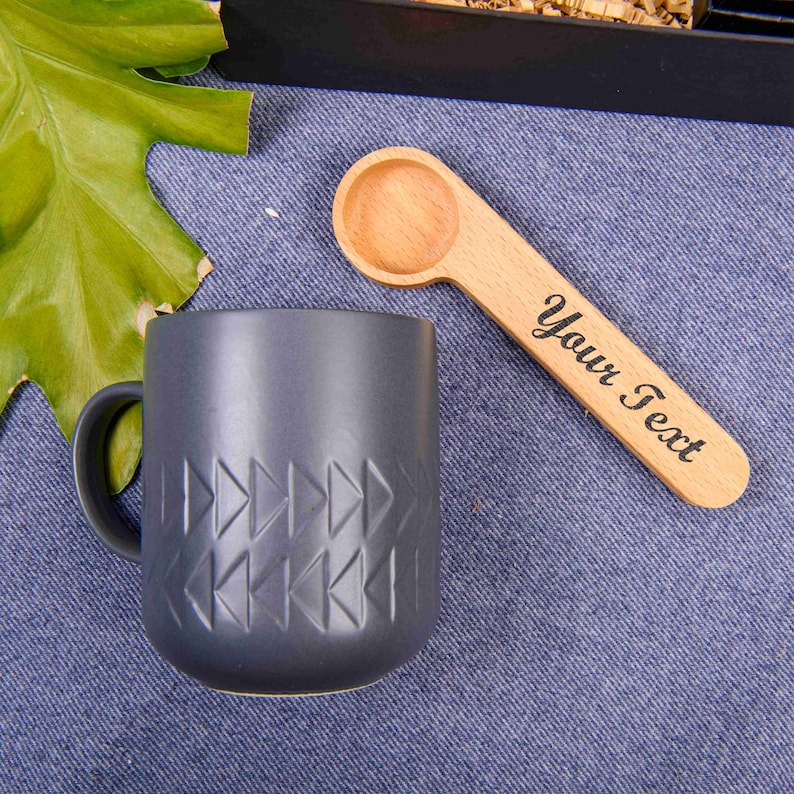 Puede incluir: Una taza de cer&aacute;mica gris oscuro con un patr&oacute;n geom&eacute;trico y una cuchara de madera con el texto "Your Text" grabado en ella.