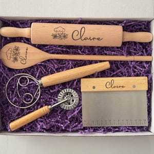 May include: A wooden baking set in a white box, including a rolling pin, spoon, whisk, dough cutter, and scraper. Each tool is engraved with the name "Claire" and floral designs. The box is filled with purple paper filler.